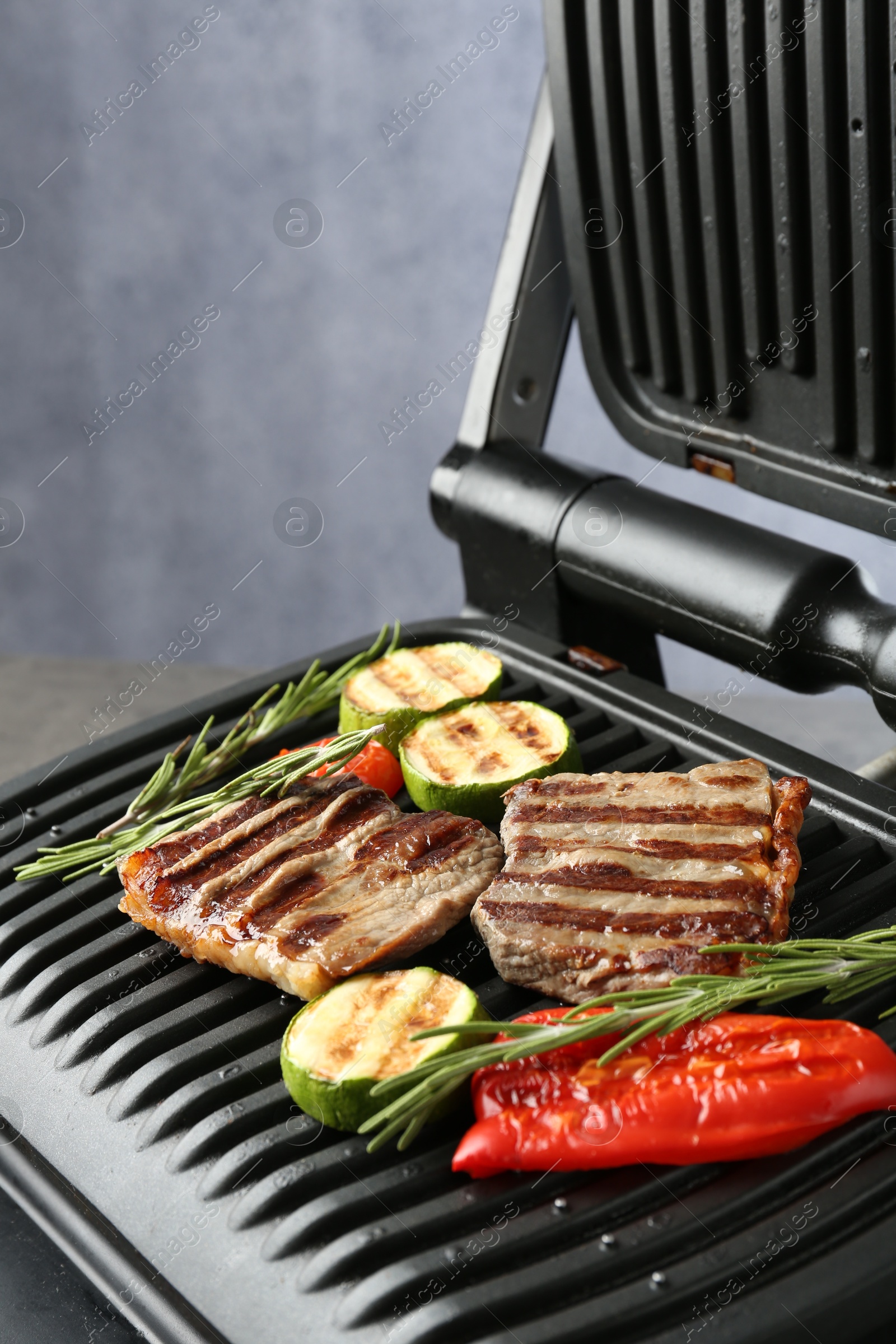 Photo of Electric grill with beef steaks, vegetables and rosemary on grey table, closeup