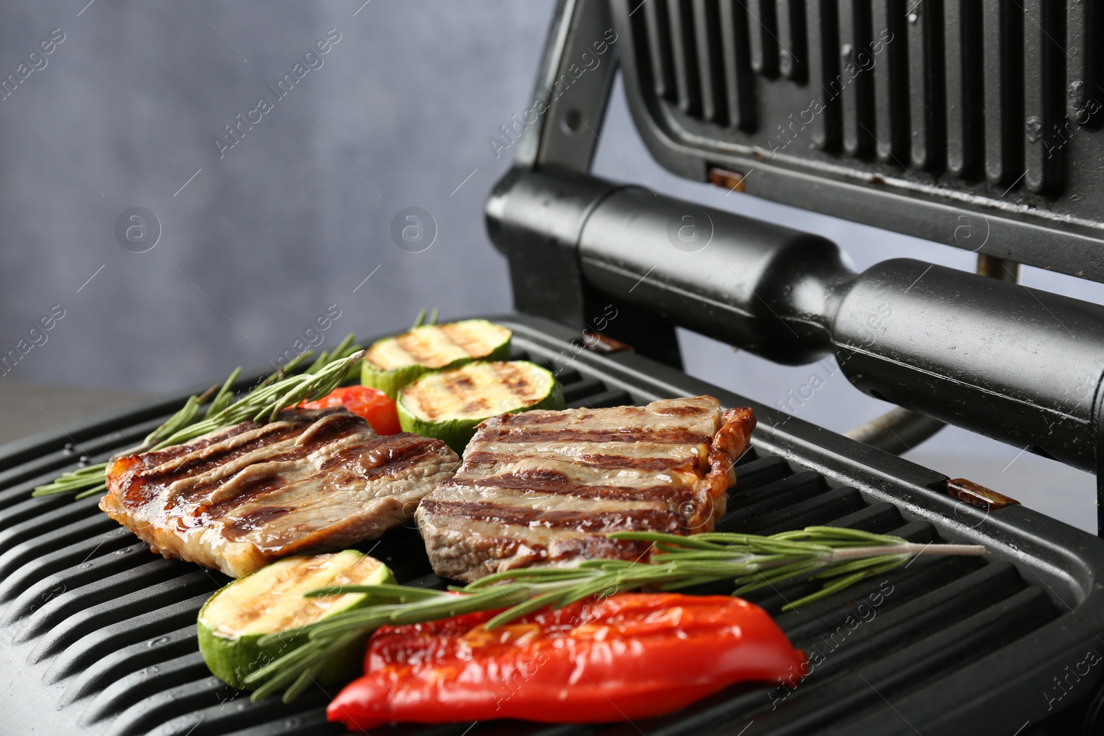Photo of Electric grill with beef steaks, vegetables and rosemary on grey table, closeup