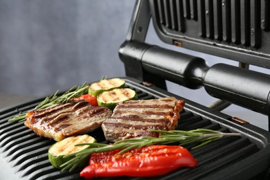Photo of Electric grill with beef steaks, vegetables and rosemary on grey table, closeup