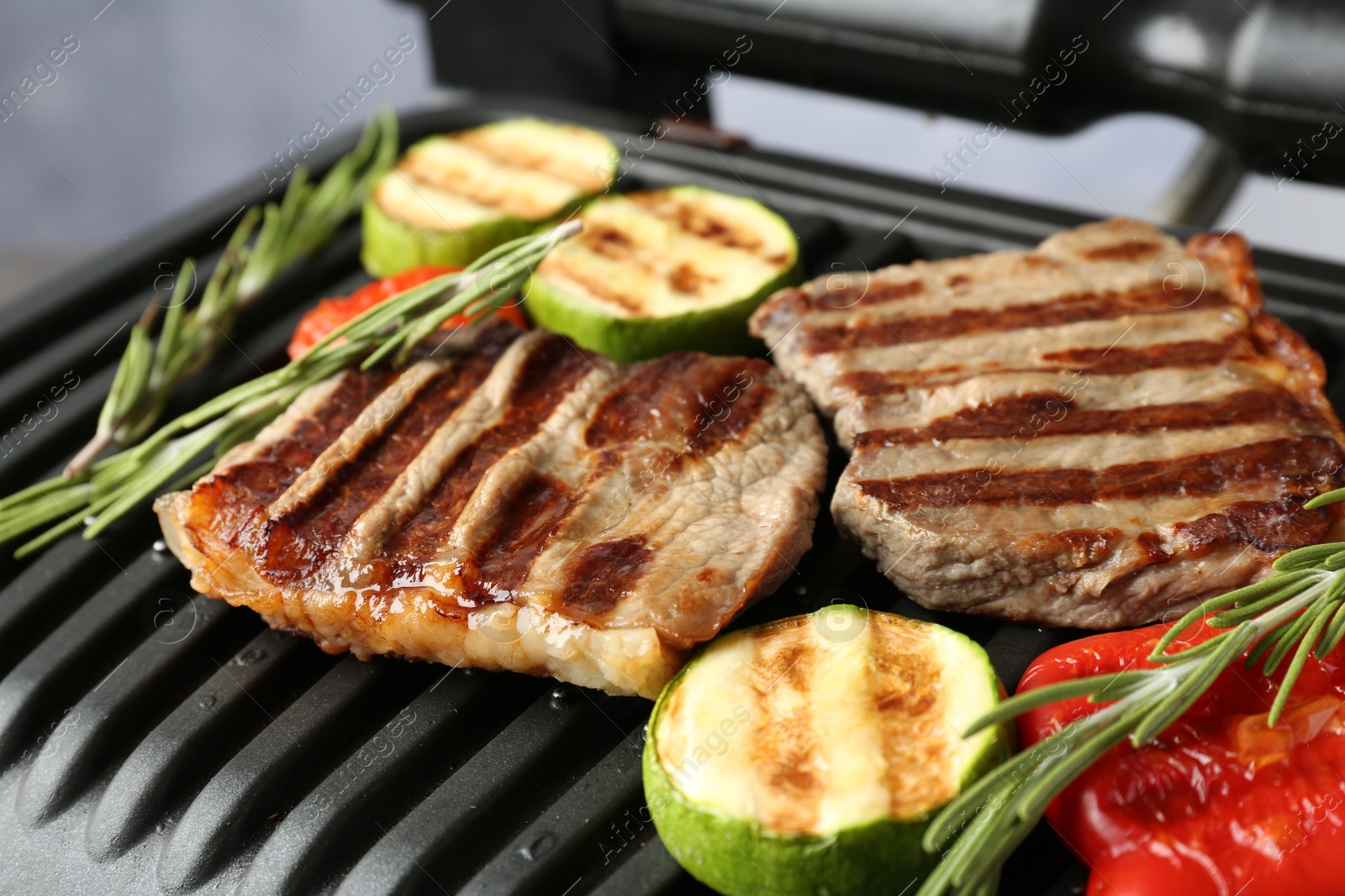 Photo of Electric grill with beef steaks, vegetables and rosemary on grey table, closeup
