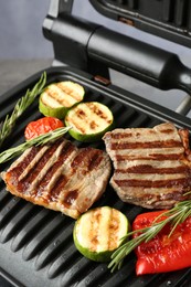 Photo of Electric grill with beef steaks, vegetables and rosemary on grey table, closeup