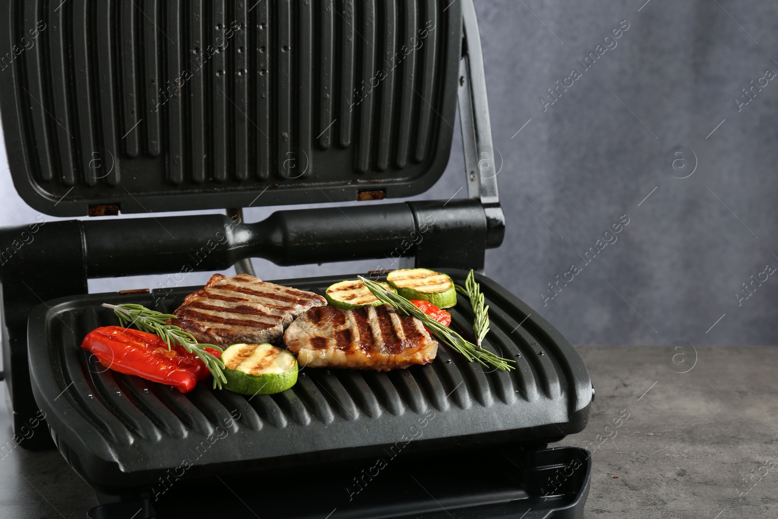Photo of Electric grill with beef steaks, vegetables and rosemary on grey table, closeup