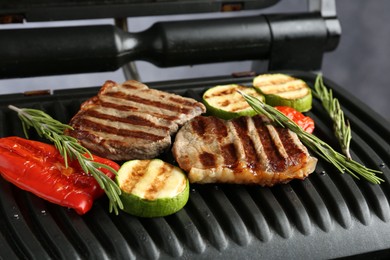 Photo of Electric grill with beef steaks, vegetables and rosemary on grey table, closeup
