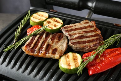 Photo of Electric grill with beef steaks, vegetables and rosemary on grey table, closeup
