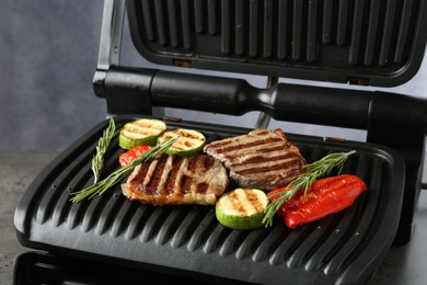 Photo of Electric grill with beef steaks, vegetables and rosemary on grey table, closeup