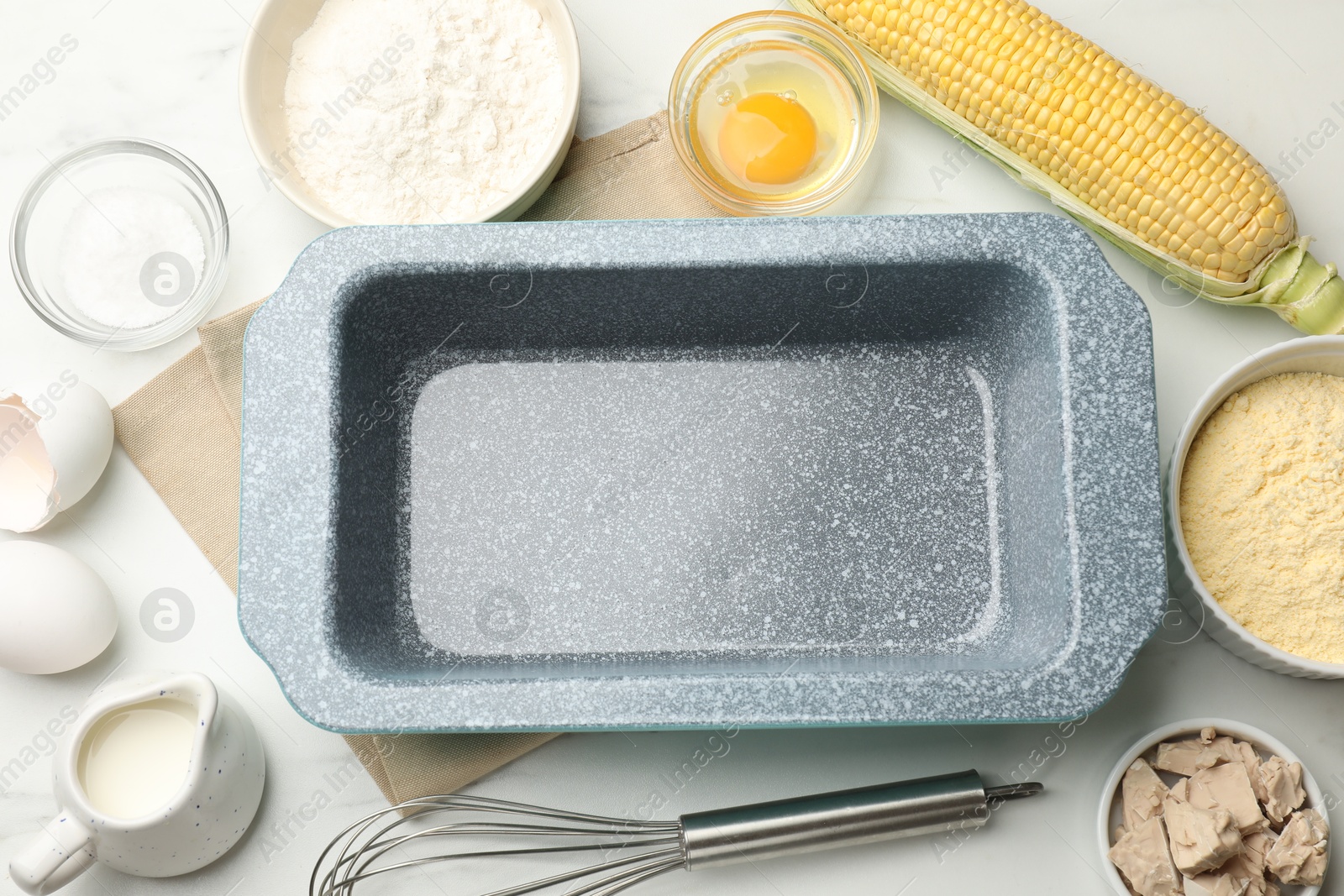 Making cornbread. Baking dish surrounded by different ingredients and whisk on white marble table, flat lay Photo of Making cornbread. Baking dish surrounded by different ingredients and whisk on white marble table, flat lay