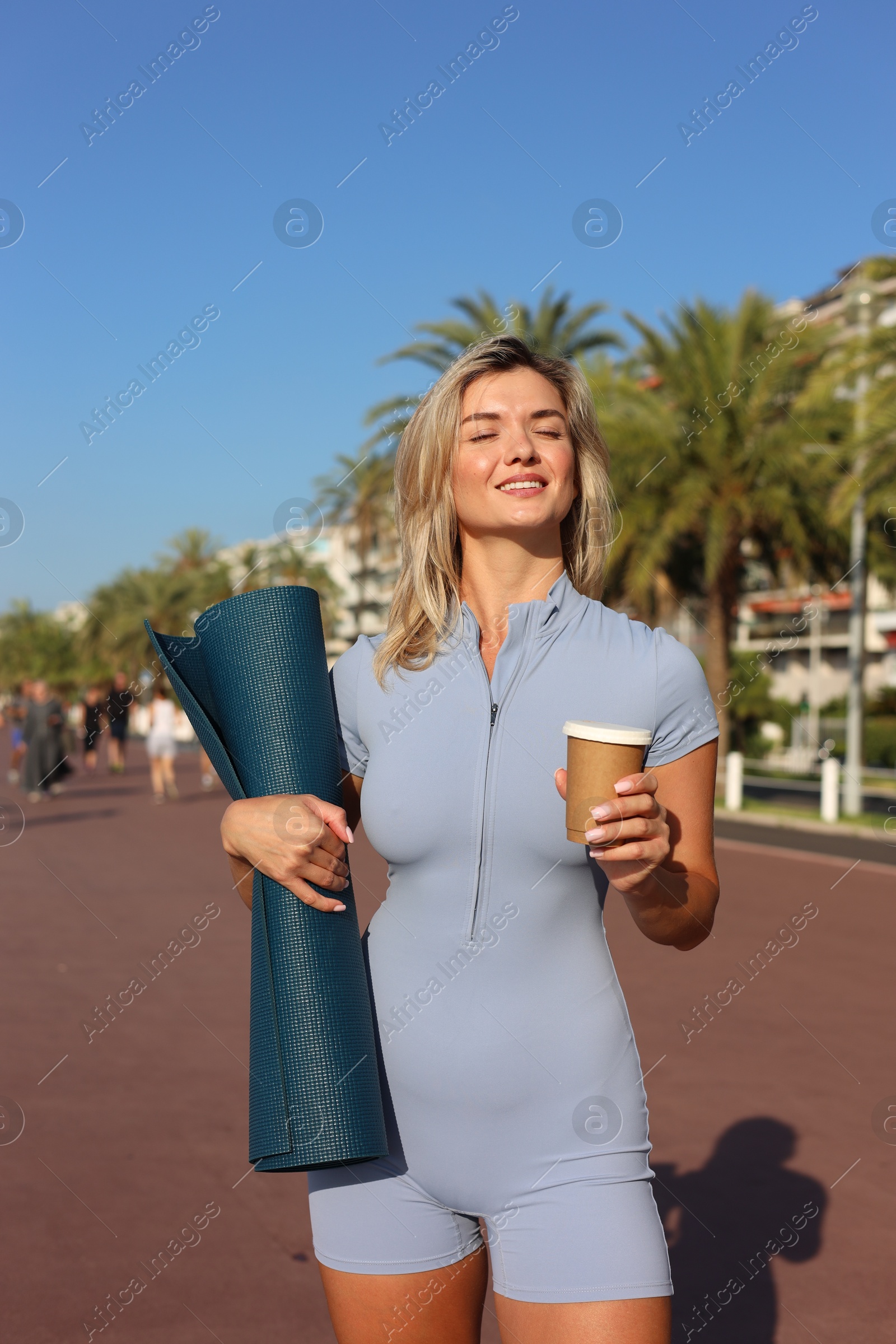 Photo of Happy young woman with yoga mat and cup of drink outdoors