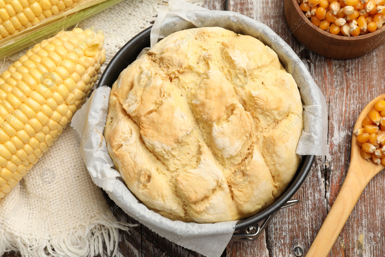 Photo of Freshly baked cornbread in cake pan, cobs and spoon on color wooden table, flat lay