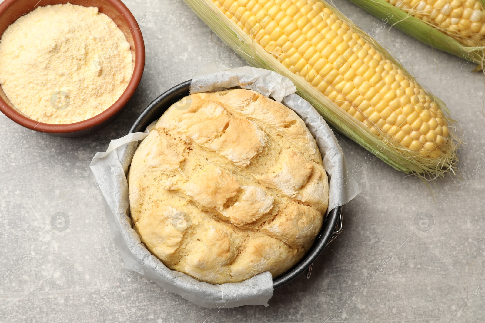 Photo of Freshly baked cornbread in cake pan, cobs and flour on grey textured table, flat lay
