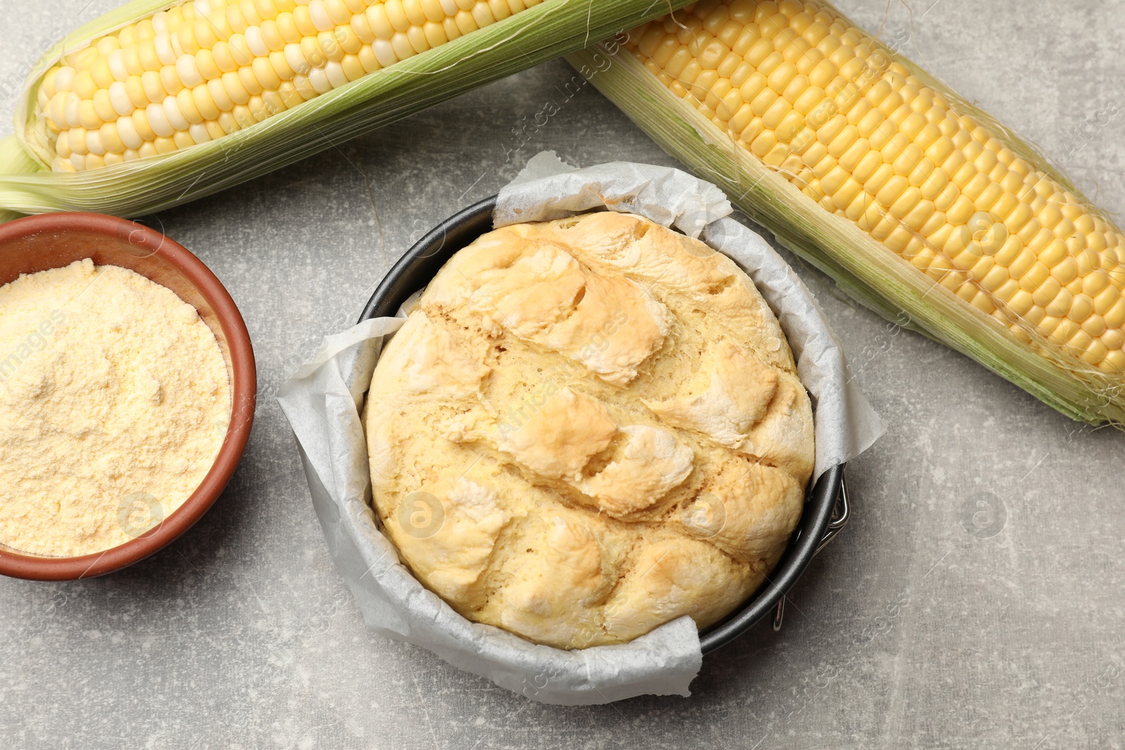 Photo of Freshly baked cornbread in cake pan, cobs and flour on grey textured table, flat lay