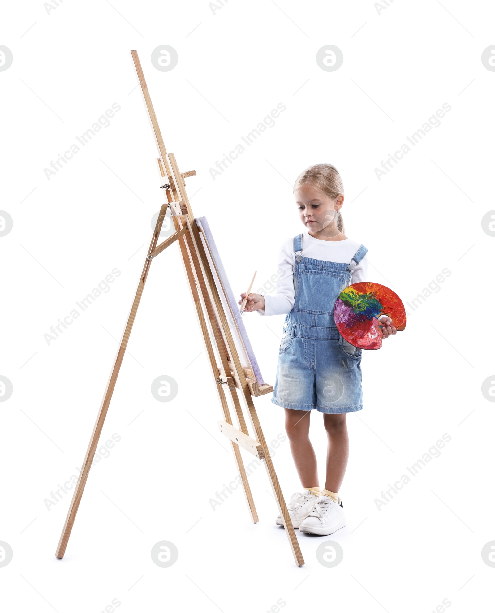 Photo of Choice of profession. Little girl with palette of paints and brush near easel pretending to be artist on white background
