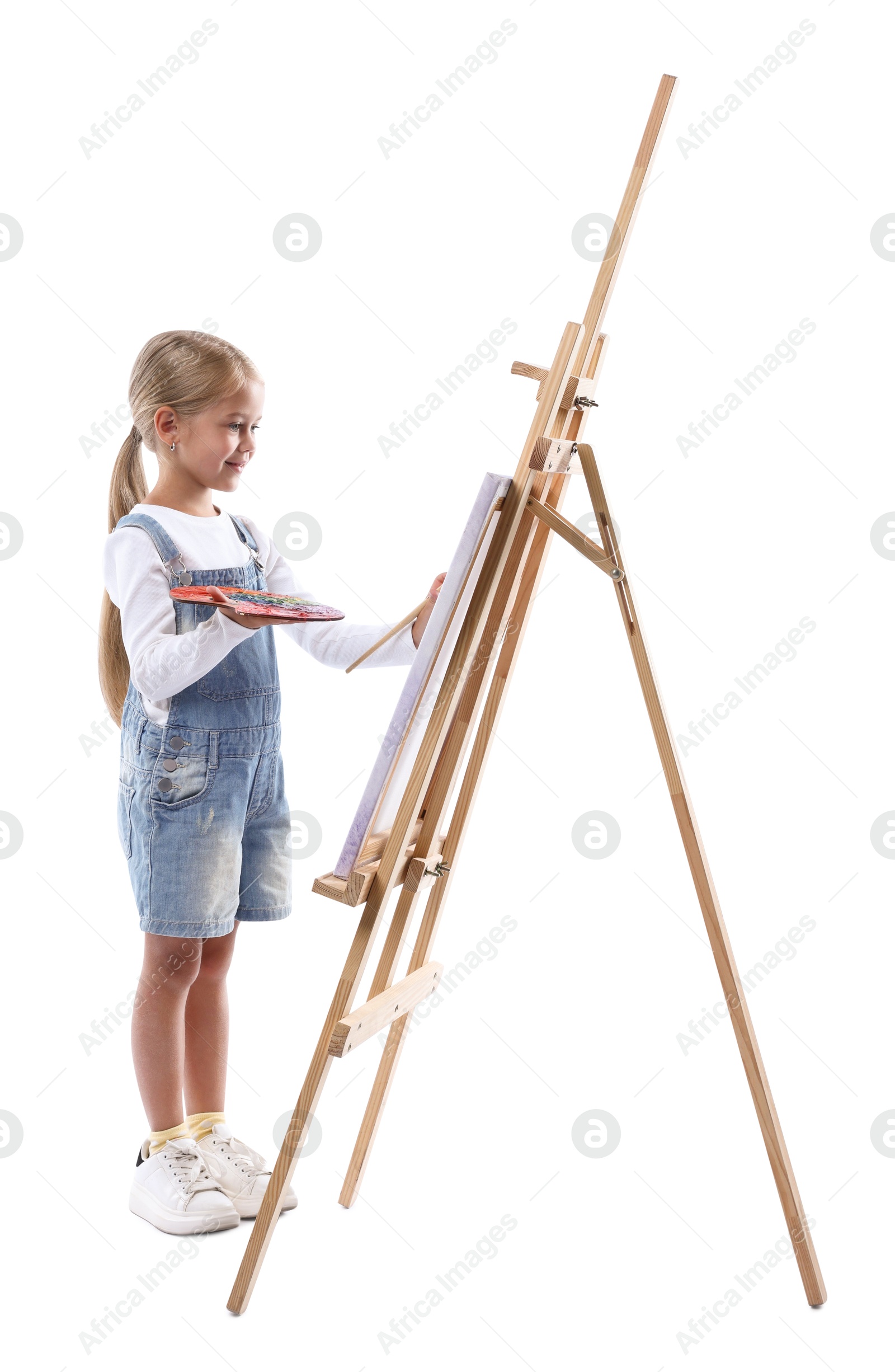 Photo of Choice of profession. Little girl with palette of paints and brush near easel pretending to be artist on white background