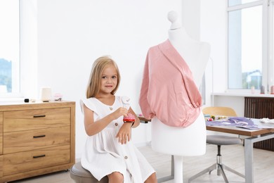 Photo of Choice of profession. Little girl with pin cushion pretending to be seamstress near mannequin in atelier