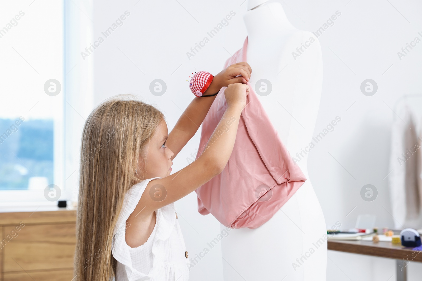 Photo of Choice of profession. Little girl with pin cushion pretending to be seamstress near mannequin in atelier