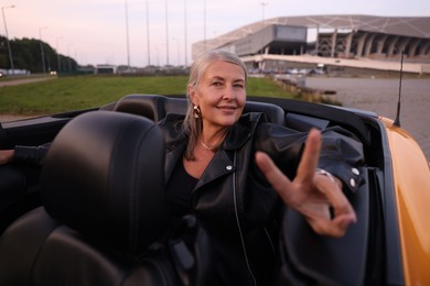 Photo of Stylish senior woman showing V-sign in modern cabriolet outdoors