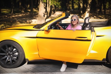 Photo of Stylish senior woman in sunglasses getting out of modern cabriolet outdoors
