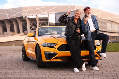 Photo of Stylish senior couple near modern cabriolet showing rock gesture outdoors