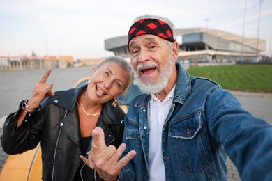 Photo of Stylish senior couple showing rock gesture while taking selfie near modern cabriolet outdoors