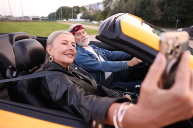 Photo of Senior woman taking selfie while man driving cabriolet outdoors, selective focus