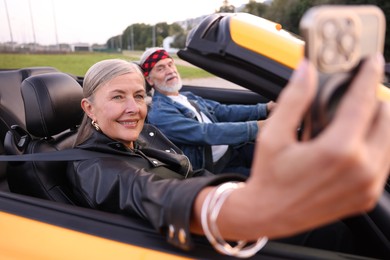 Photo of Senior woman taking selfie while man driving cabriolet outdoors, selective focus