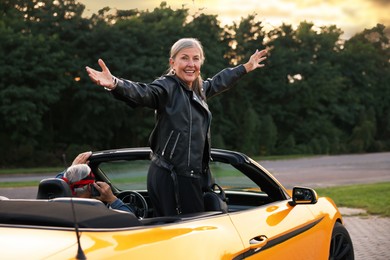 Photo of Senior couple in modern cabriolet outdoors. Woman with wide open arms and man talking on phone