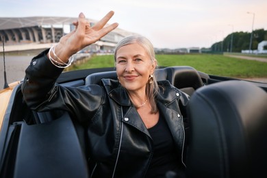 Photo of Stylish senior woman showing V-sign in modern cabriolet outdoors