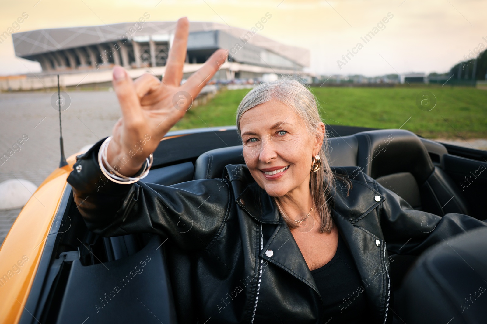 Photo of Stylish senior woman showing V-sign in modern cabriolet outdoors