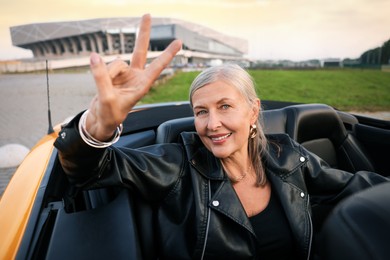 Photo of Stylish senior woman showing V-sign in modern cabriolet outdoors