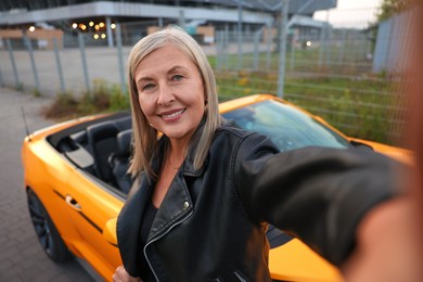 Photo of Smiling senior woman taking selfie near modern cabriolet outdoors