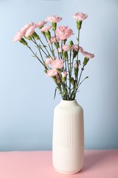 Photo of Beautiful carnation flowers in vase on pink table against light blue background