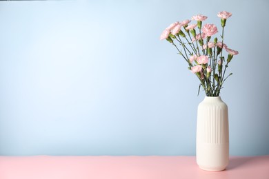 Photo of Beautiful carnation flowers in vase on pink table against light blue background, space for text
