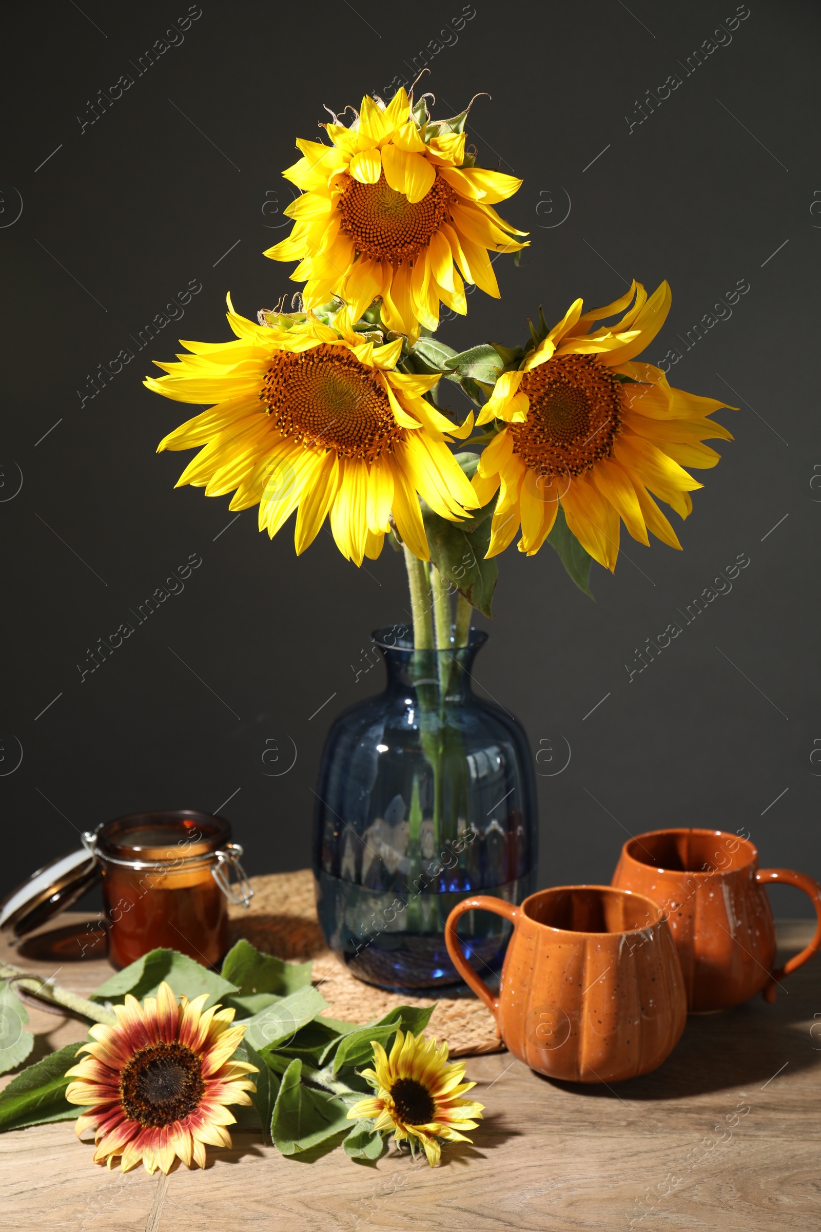 Beautiful sunflowers in vase, candle and cups on wooden table against black background Photo of Beautiful sunflowers in vase, candle and cups on wooden table against black background