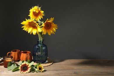Photo of Beautiful sunflowers in vase, candle and cups on wooden table against black background, space for text