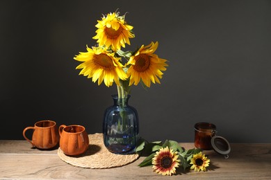Photo of Beautiful sunflowers in vase, candle and cups on wooden table against black background