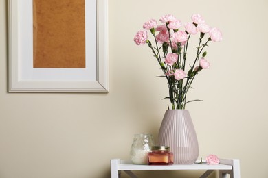 Photo of Beautiful carnation flowers in vase and candles on white stand near beige wall indoors