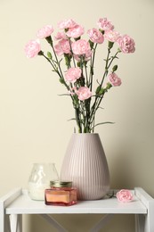 Photo of Beautiful carnation flowers in vase and candles on white stand near beige wall indoors