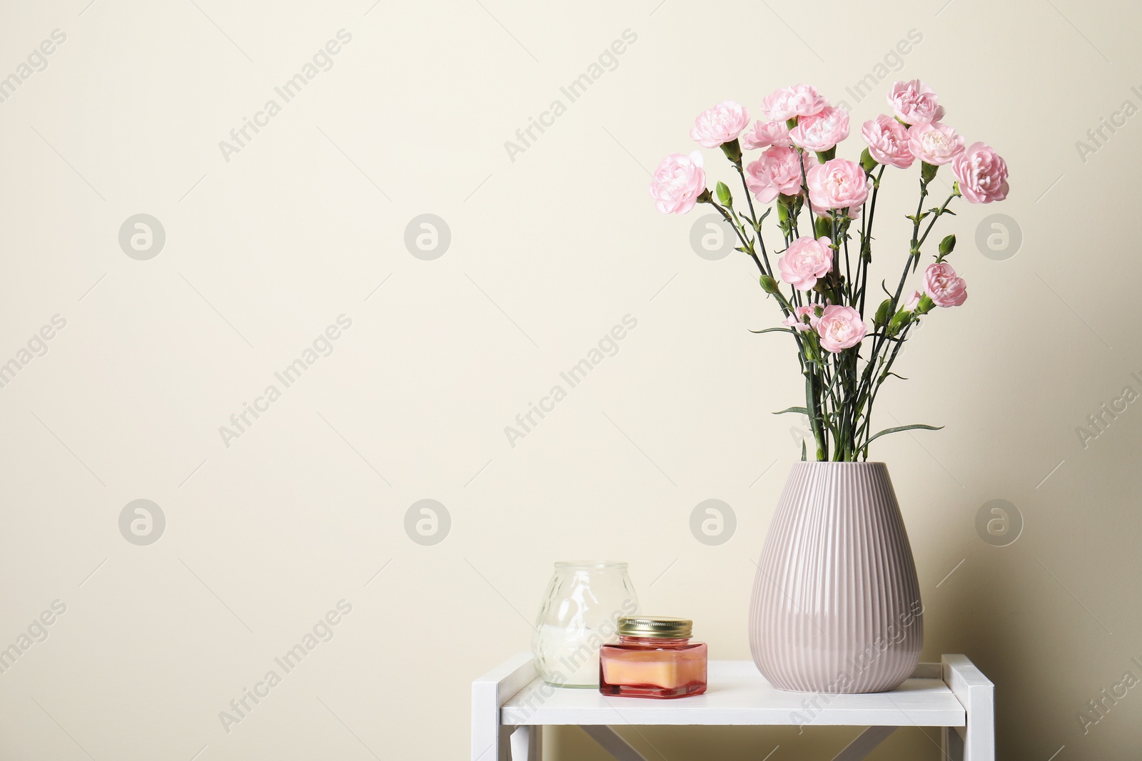 Beautiful carnation flowers in vase and candles on white stand near beige wall indoors, space for text Photo of Beautiful carnation flowers in vase and candles on white stand near beige wall indoors, space for text
