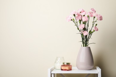 Photo of Beautiful carnation flowers in vase and candles on white stand near beige wall indoors, space for text
