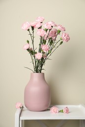 Photo of Beautiful carnation flowers in vase on white stand near beige wall indoors