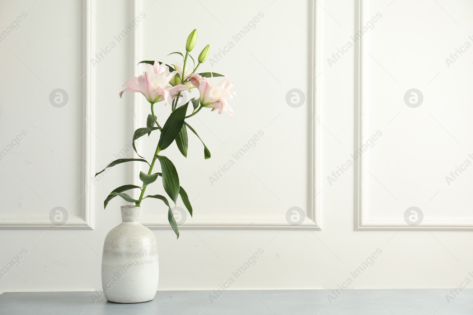 Beautiful lily flowers in vase on grey table near white wall, space for text Photo of Beautiful lily flowers in vase on grey table near white wall, space for text