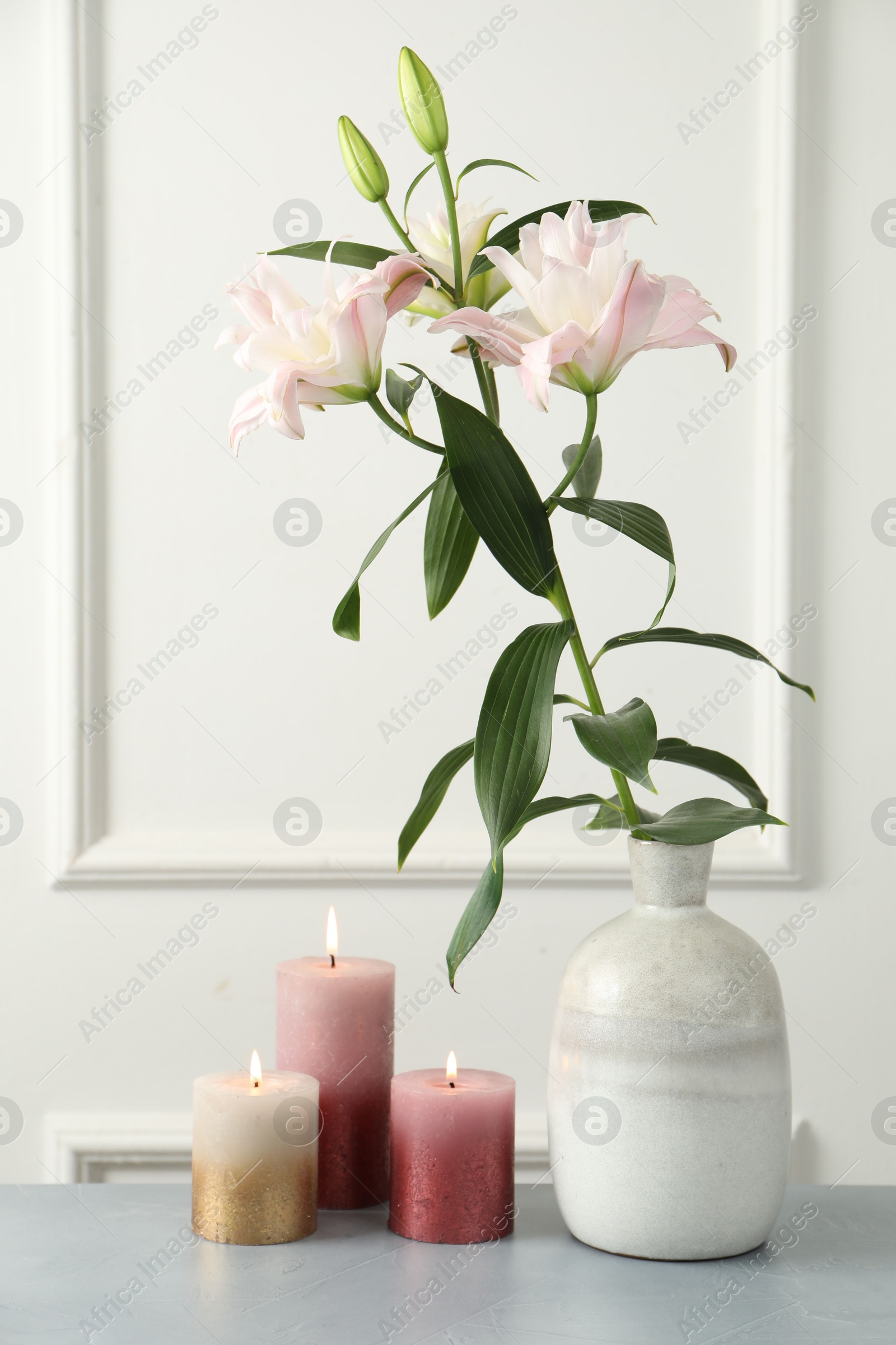 Photo of Beautiful lily flowers in vase and burning candles on grey table near white wall indoors