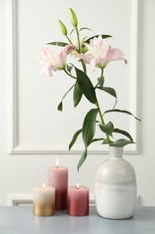 Photo of Beautiful lily flowers in vase and burning candles on grey table near white wall indoors