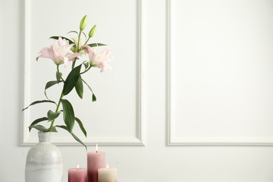 Photo of Beautiful lily flowers in vase and burning candles near white wall indoors, space for text