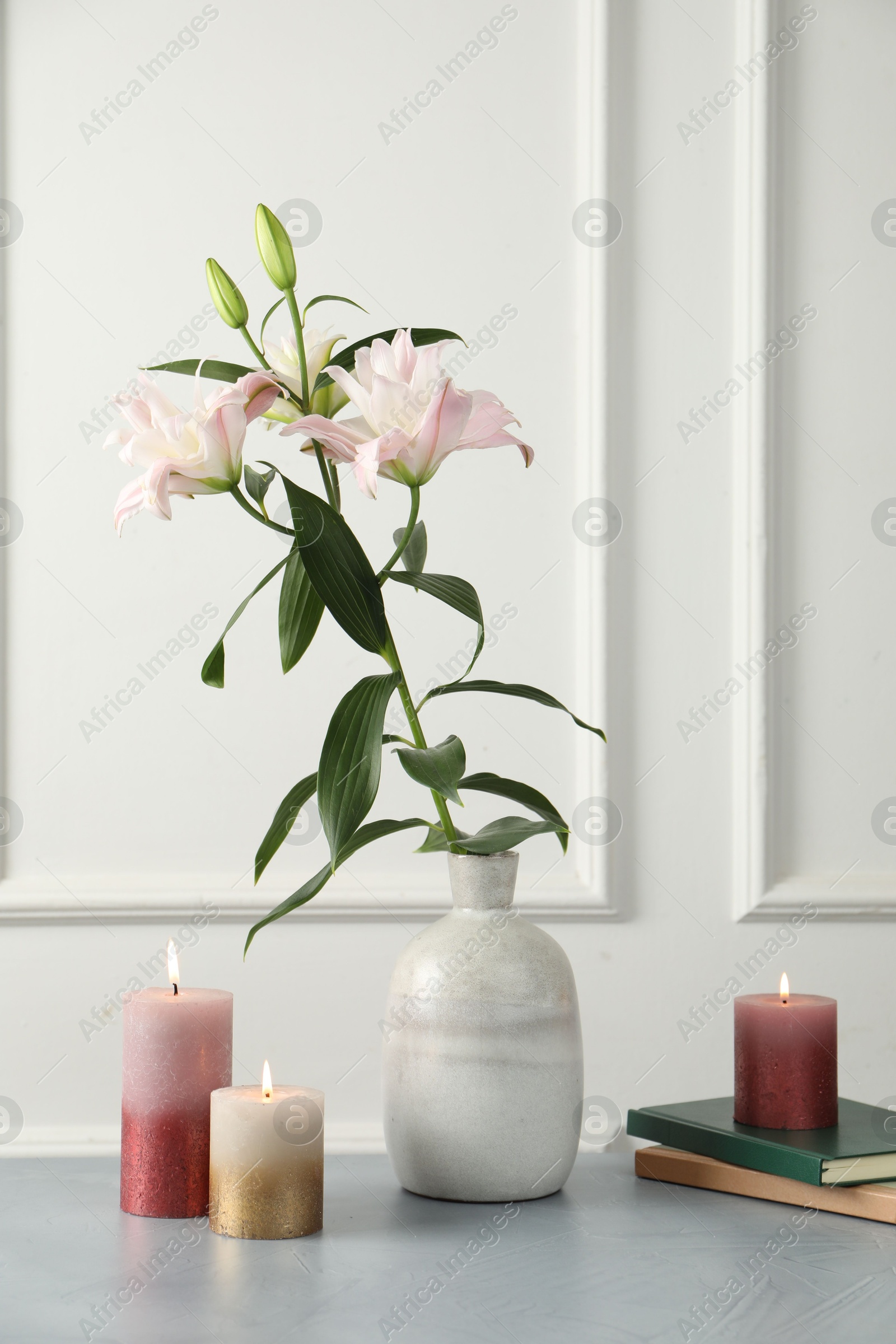 Photo of Beautiful lily flowers in vase, books and burning candles on grey table near white wall indoors