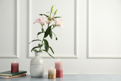 Photo of Beautiful lily flowers in vase, books and burning candles on grey table near white wall indoors, space for text