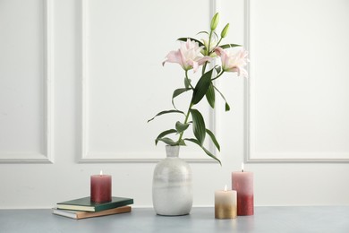 Photo of Beautiful lily flowers in vase, books and burning candles on grey table near white wall indoors
