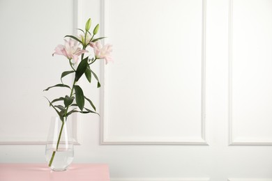 Photo of Beautiful lily flowers in glass vase on pink wooden table near white wall indoors, space for text