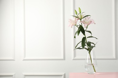 Photo of Beautiful lily flowers in glass vase on pink wooden table near white wall indoors, space for text