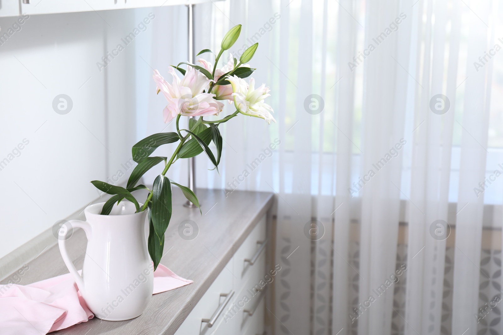 Beautiful lily flowers in jug on wooden cabinet indoors, space for text Photo of Beautiful lily flowers in jug on wooden cabinet indoors, space for text