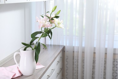 Photo of Beautiful lily flowers in jug on wooden cabinet indoors, space for text
